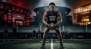 A hyper-realistic action portrait of a dominant high school defensive tackle exploding off the line during a spring practice in Georgia, dirt flying from cleats, offensive lineman resistance visible in background blur, powerful athletic stance with extreme muscle definition and focus, stormy sky over rural high school stadium, cinematic lighting with high contrast shadows, ESPN recruiting feature photography style, deep greens, steel gray clouds, and warm sunset highlights, no branding, raw intensity emphasizing elite SEC-level talent evaluation, Gemini Nano Banana optimized